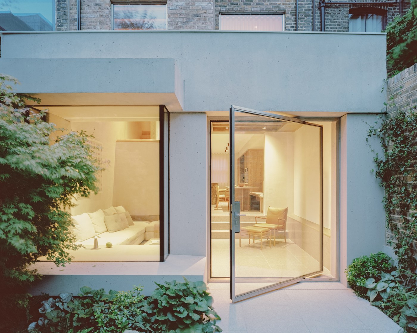 Caithness Road W14 — concrete rear facade at dusk, with a large fixed window and a full-height stainless-steel pivot door open onto the garden